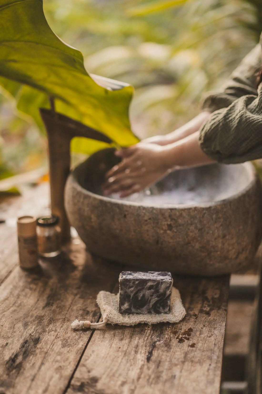 Natural soap bar on rustic soap dish beside stone basin with hands washing, surrounded by greenery