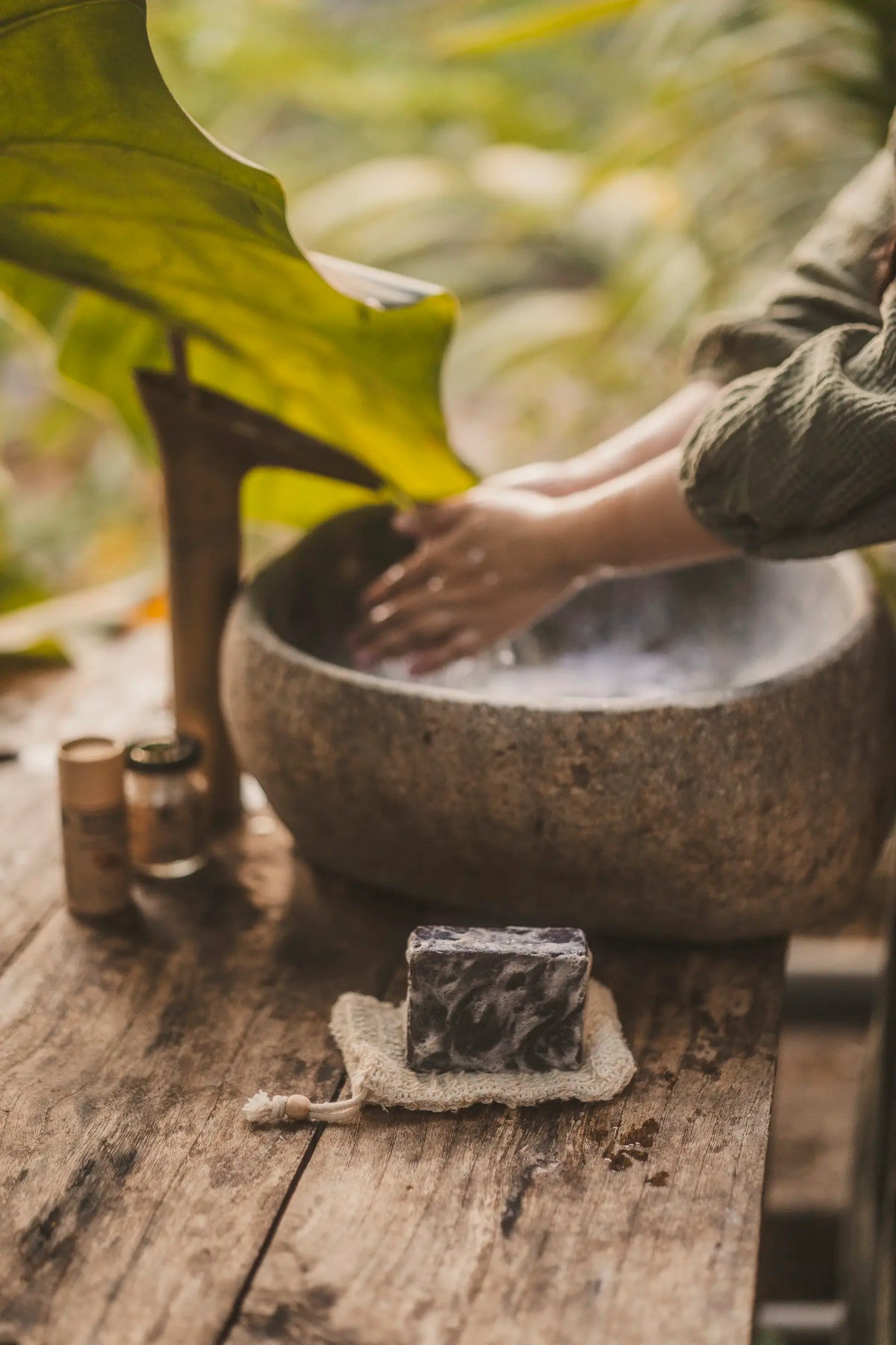 Natural soap bar on rustic soap dish beside stone basin with hands washing, surrounded by greenery