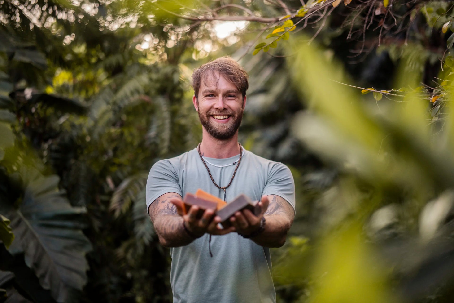 Man holding natural soap bars outdoors in lush greenery, smiling for adventure-ready clean skincare